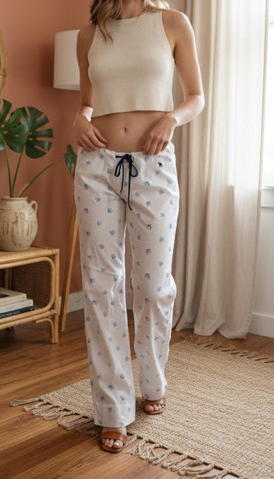 Woman in a white top and patterned pants standing in a room with a pink wall, wooden floor, and a window with curtains.