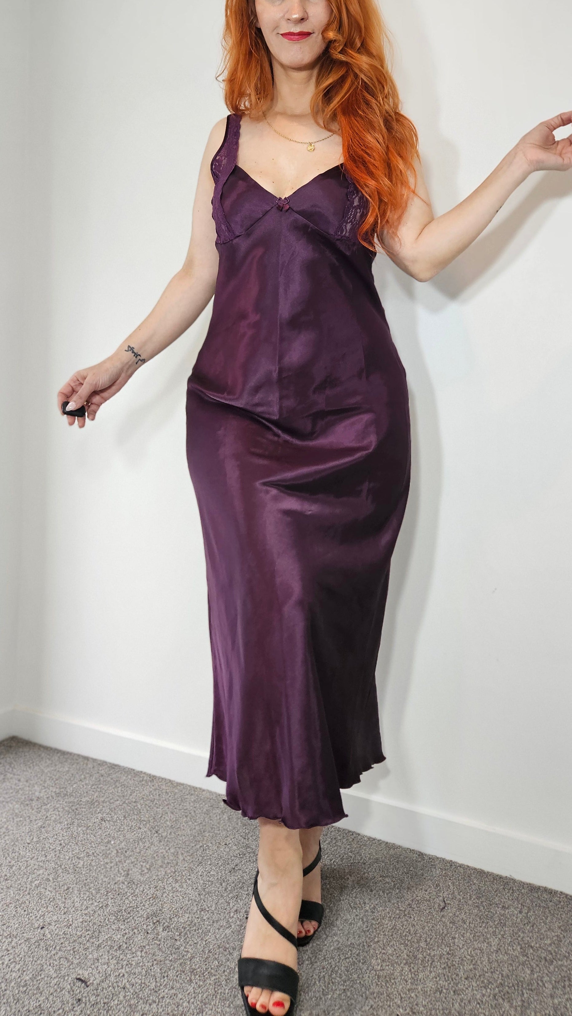 Deep plum purple satin-finish slip dress with lace-trimmed neckline, thin straps and flowing long skirt, modelled on a woman.
